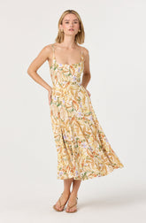 A woman is wearing a sleeveless, ankle-length dress with a fitted bodice and thin spaghetti straps. The dress features a light background with a colorful floral and leaf pattern in shades of orange, green, purple, and beige. She is also wearing beige open-toe high-heeled sandals.